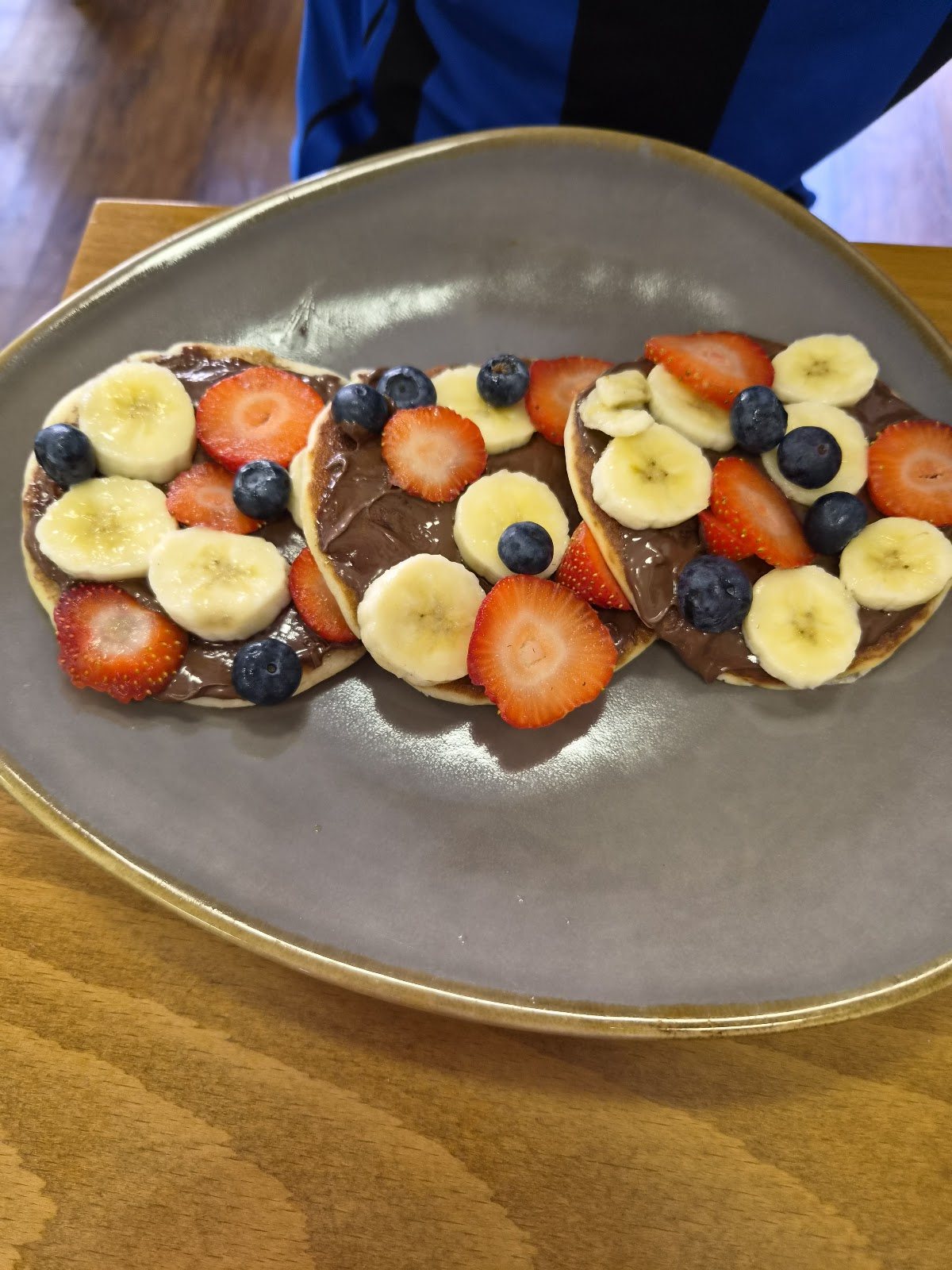 Three pancakes spread with chocolate and topped with sliced banana, strawberries and blueberries on a grey plate.