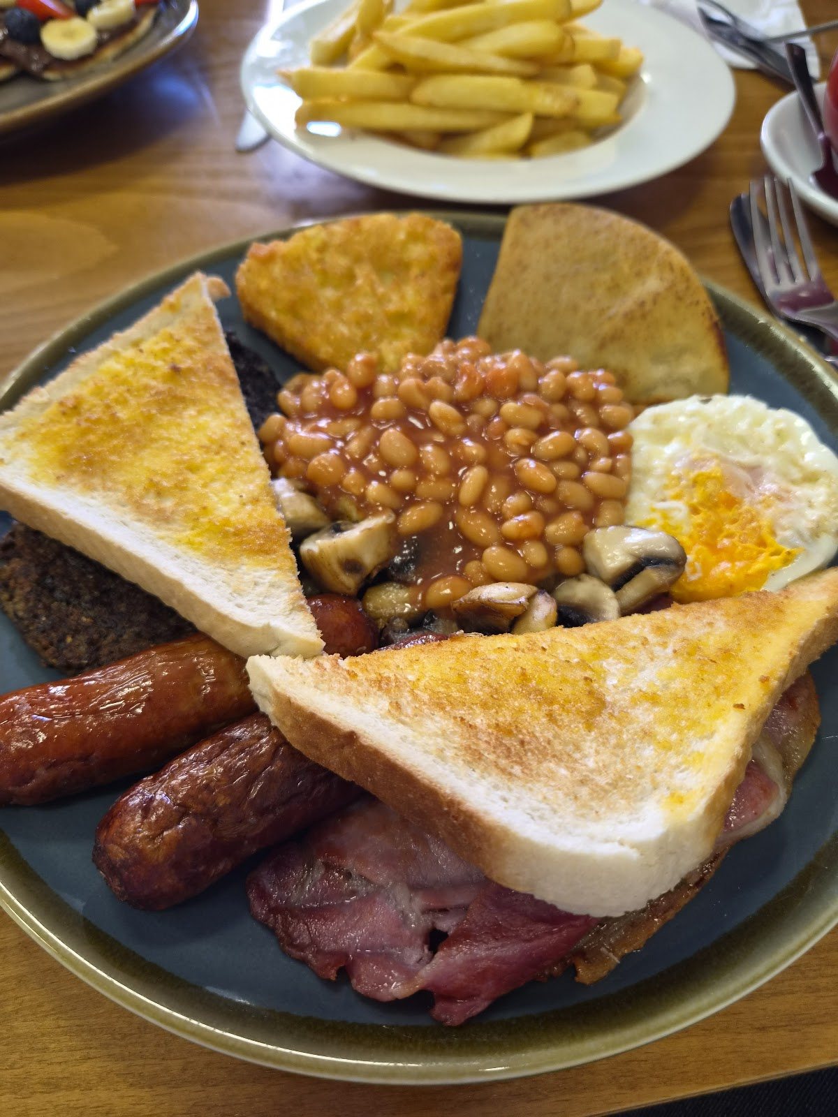 A big full breakfast on a slate-blue plate: two sausages, bacon, a fried egg, beans with mushrooms, black pudding, a hash brown, a tattie scone and buttered toast, with chips on the side.