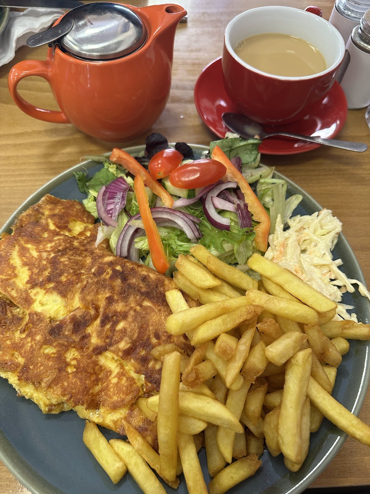 A golden omelette with chips, a side salad of tomatoes, red onion, peppers and olives, and coleslaw, next to an orange teapot and a red cup of tea.