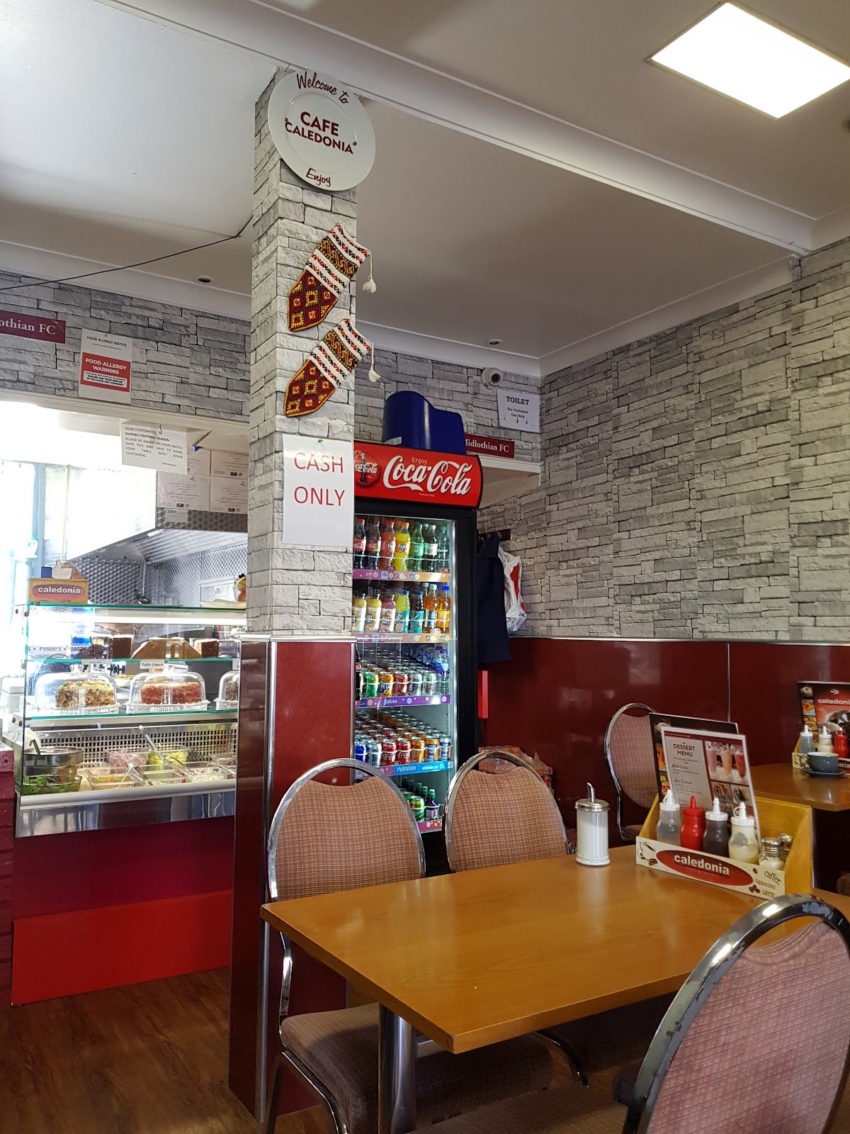 Inside Caledonia: a pillar with a 'Welcome to Cafe Caledonia' sign, stone-effect walls, maroon panelling, wooden tables with upholstered chairs, a chiller cabinet of cakes and a drinks fridge.